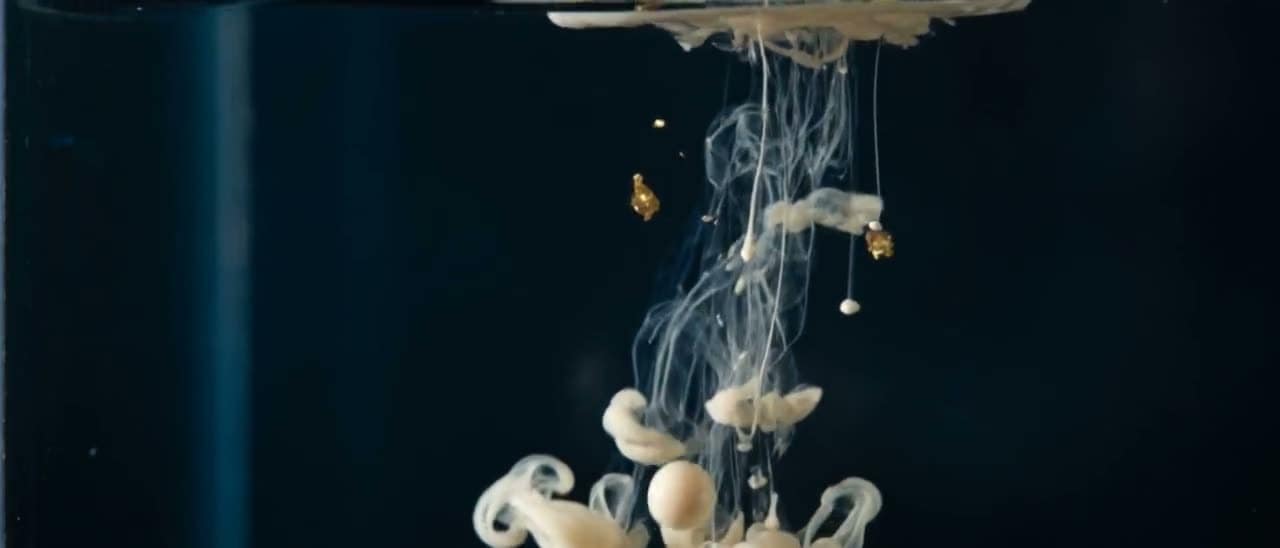A drop of cream ink releasing from the surface of dark water and falling, thin tendrils trailing behind it.