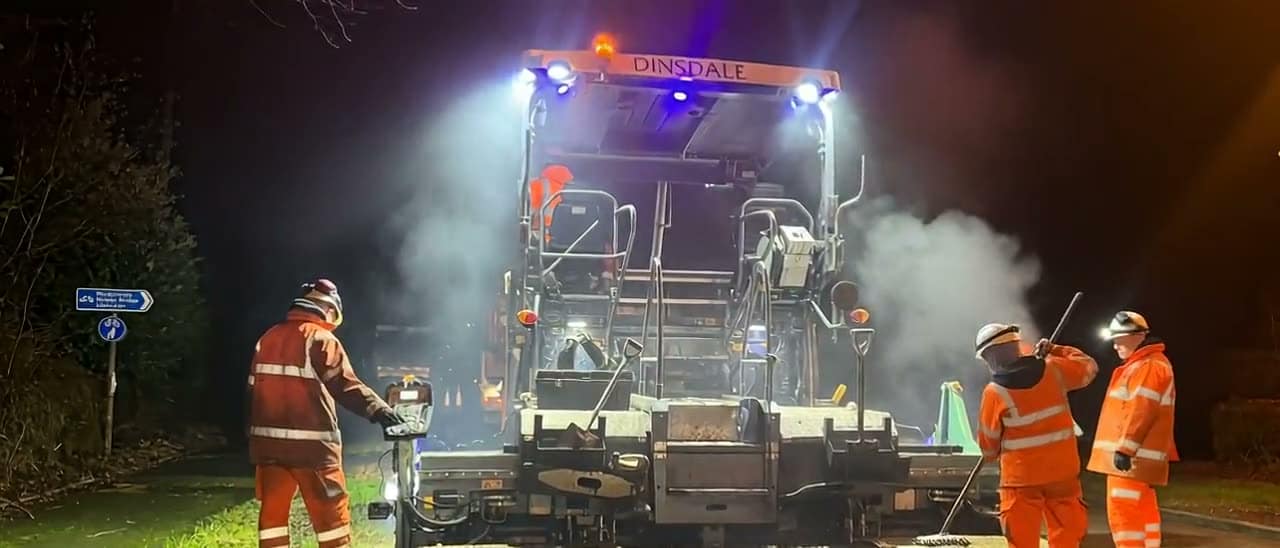 A Dinsdale Contracts paver working under floodlights at night, steam rising, crew alongside in high visibility gear.